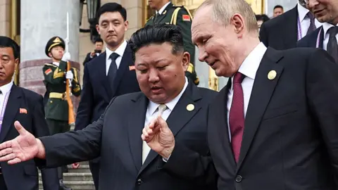 Getty Images Russian President Vladimir Putin and North Korean leader Kim Jong Un attend a meeting in Beijing. They are walking down some stairs and talking to each other. Bodyguards and other officials lurk in the background.