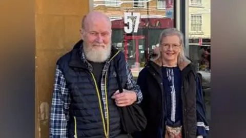 Family handout Peter on the right of this image is shown after a recent hair cut back in the UK. His beard is trimmed and his hair shorter. His wife, Barbie, stands smiling next to him.