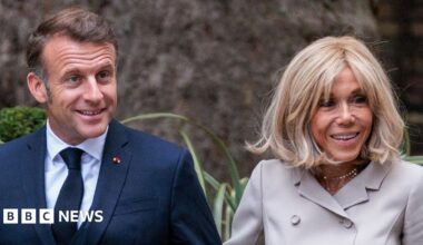 French President Emmanuel Macron and First Lady Brigitte Macron arriving in Downing Street in July. The image depicts the couple walking arm in arm. Mr Macron is dressed in a dark blue suit, white shirt, and dark tie. Mrs Macron is wearing a light-colored, double-breasted jacket suit. They are surrounded by greenery, including bushes and plants with long leaves