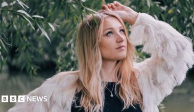 Dolly Mavies next to a tree, the leaves touching her head. She has long fair hair and blue eyes. She wears a black top with a white, feathery jacket.