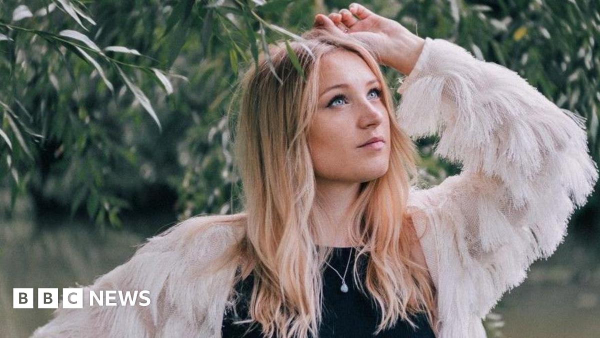 Dolly Mavies next to a tree, the leaves touching her head. She has long fair hair and blue eyes. She wears a black top with a white, feathery jacket.