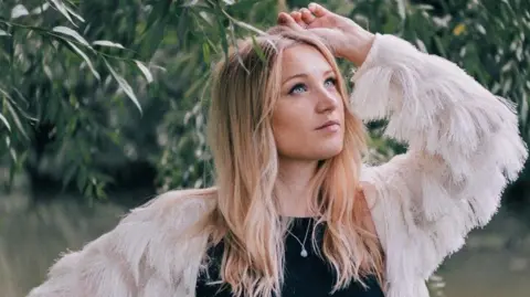 Tess Viera Photography Dolly Mavies next to a tree, the leaves touching her head. She has long fair hair and blue eyes. She wears a black top with a white, feathery jacket.