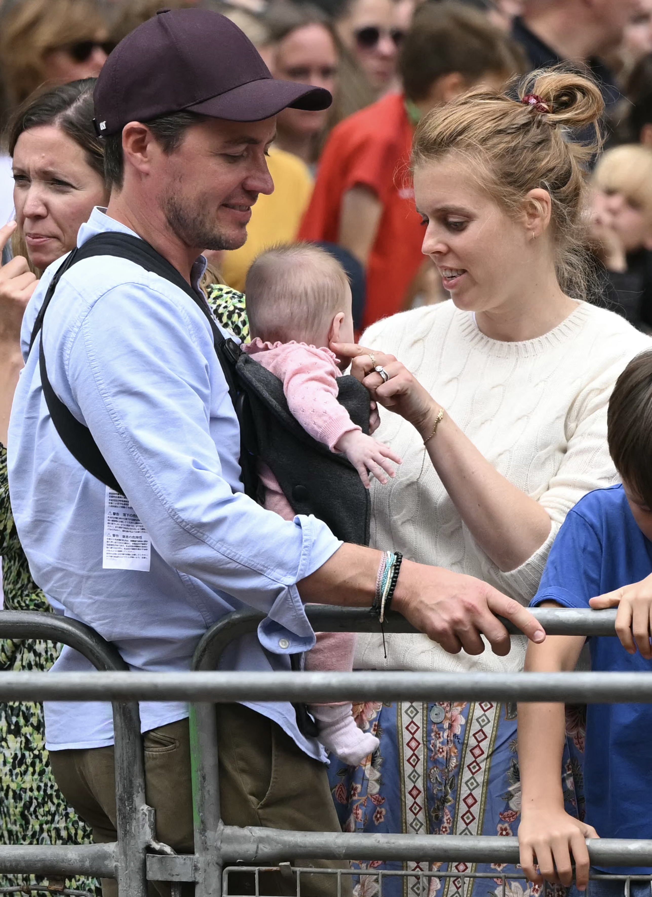 Princess Beatrice wearing a cream sweater talking to baby Athena, who is wearing a pink outfit and sitting in a carrier on Edoardo Mapelli Mozzi's chest