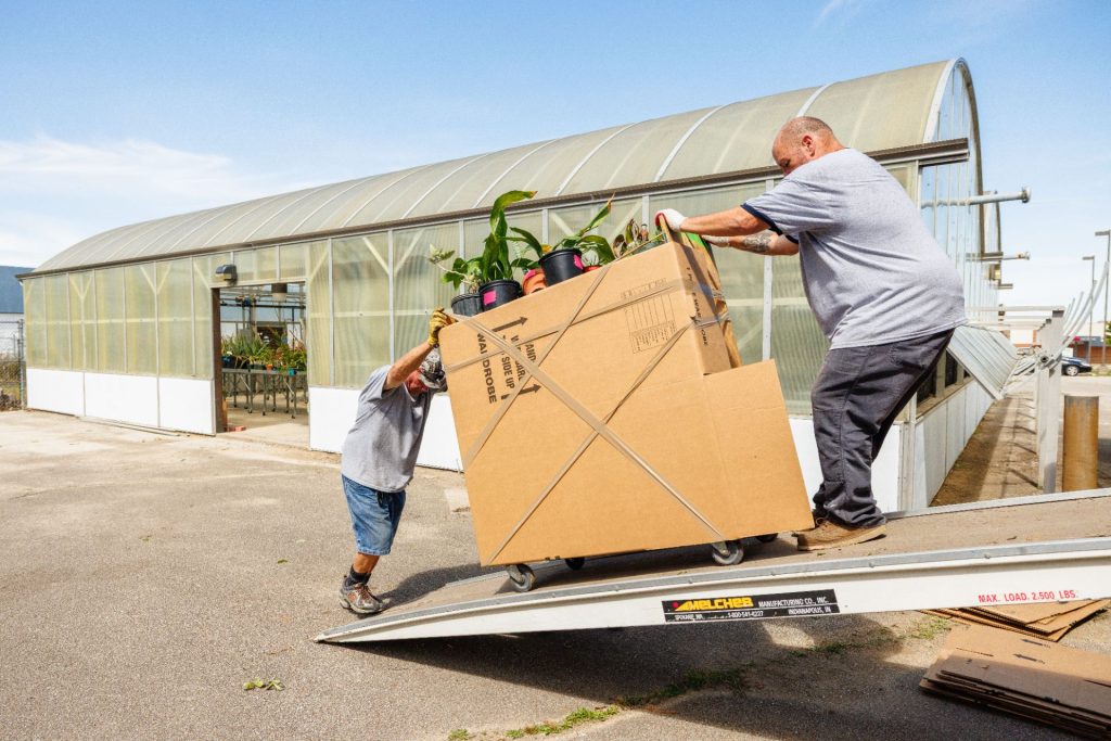 movers push and pull a box filled with plants down a ramp from a moving truck
