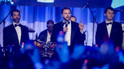 Getty Images Three men in tuxedos in a line, Trump Jr on the right, Vance in the centre holding a microphone and speaking and Kirk on the right. There is a band in the background and many people are holding up phones to take pictures in the foreground