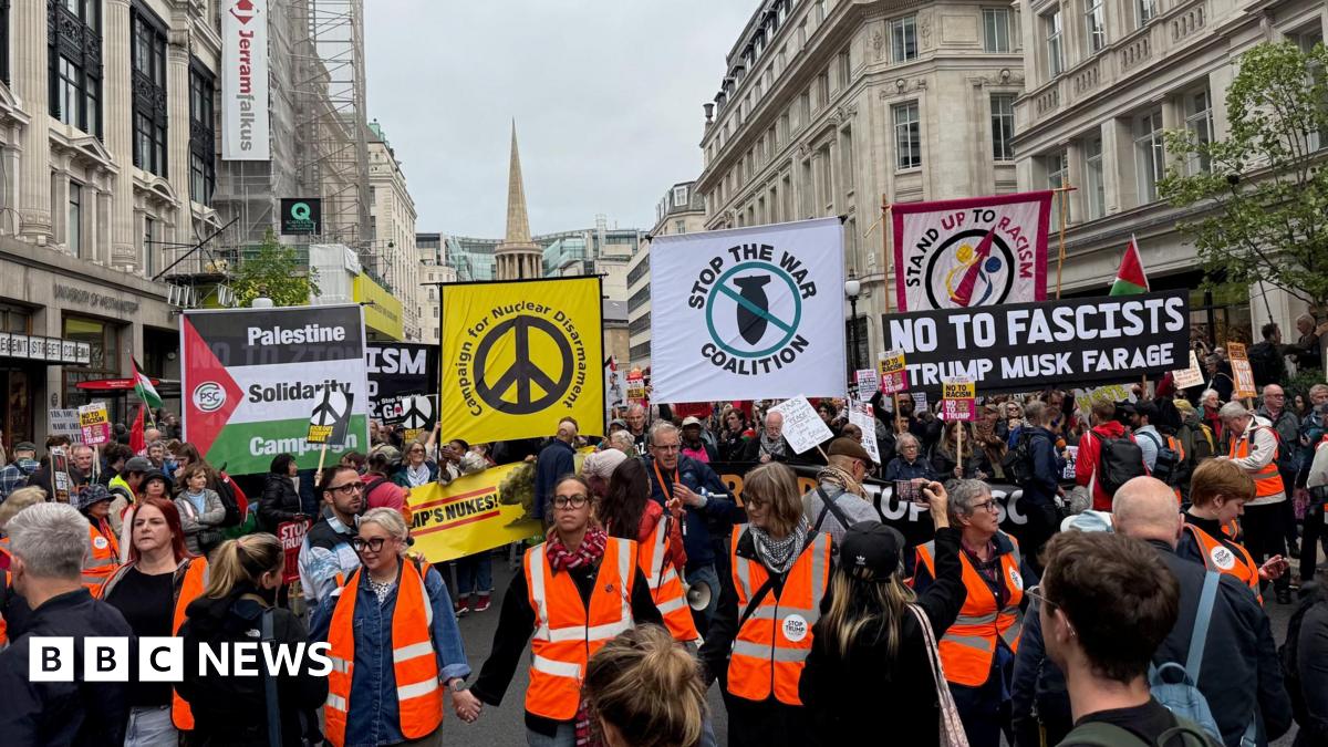 People wearing orange reflective jackets holding hands at the front of the march. Behind them are crowds of people some holding banners, including some that read: Palestine Solidarity Campaign, Campaign for Nuclear Disarmament, Stop the War Coalition, No to Fascists, and Stand Up to Racism.