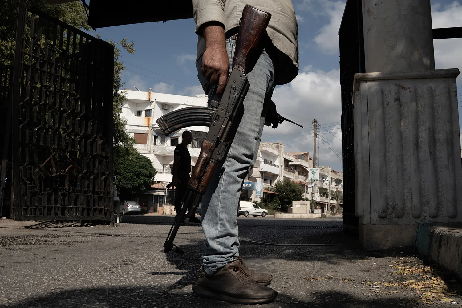 A man, seen from the waist down, holds a gun at his side. Behind him is a gate and a four-story building and street.