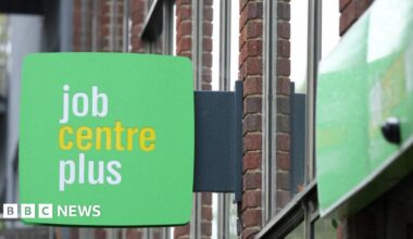 A close up of a green sign hanging off a building that reads "job centre plus". The word "centre" is in yellow and the other two words are in white.