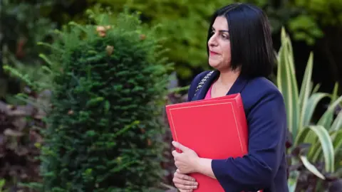 PA Media Home Secretary Shabana Mahmood walks outside Downing Street, holding a large red folder. She wears a pink dress, a navy blue blazer, and black shoes. Behind her is a well-kept garden with vibrant flowers and green shrubs.