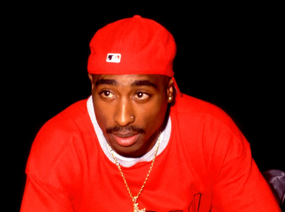 Tupac Shakur wearing a red cap and sweater, with a gold chain, against a black background.