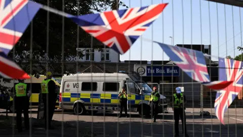 Reuters Police officers wearing hi-vis jackets guard The Bell Hotel in Epping. A white police van blocks the entrance, as does a long line of fences. Union Jack flags have been tied to one of the fences.