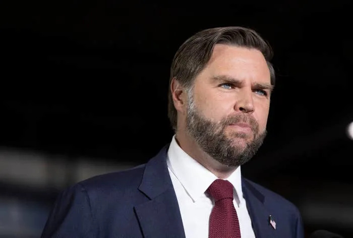 A person in a suit and tie looks serious, facing slightly to the side. The background is blurred