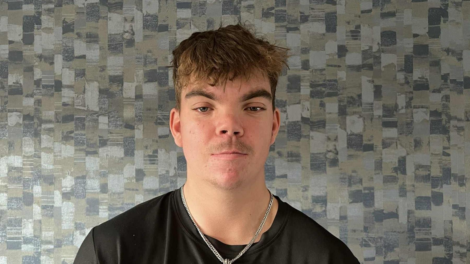 The picture shows a man wearing a black t-shirt with a silver chain around his neck. The man is standing in front of patterned wallpaper