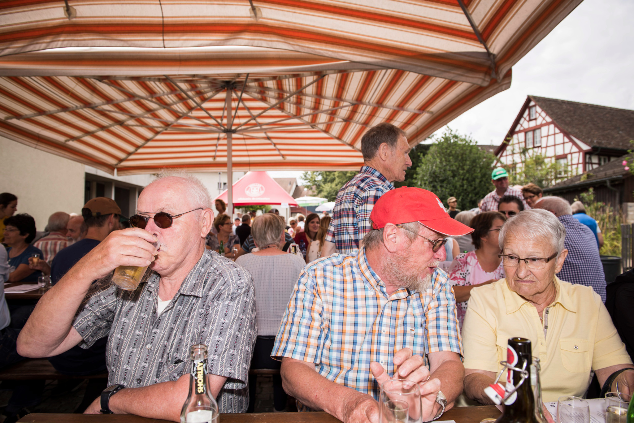 Zahlreiche Menschen sitzen unter rot gestreiften Markisen bei der 1. August Feier in Dietlikon ZH, darunter auch ältere Herrschaften, die Bier trinken.