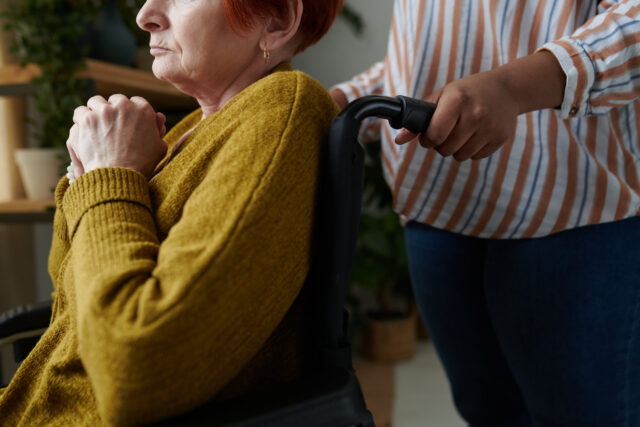 Close-up of senior woman with disability sitting in wheelchair with caregiver standing behind her