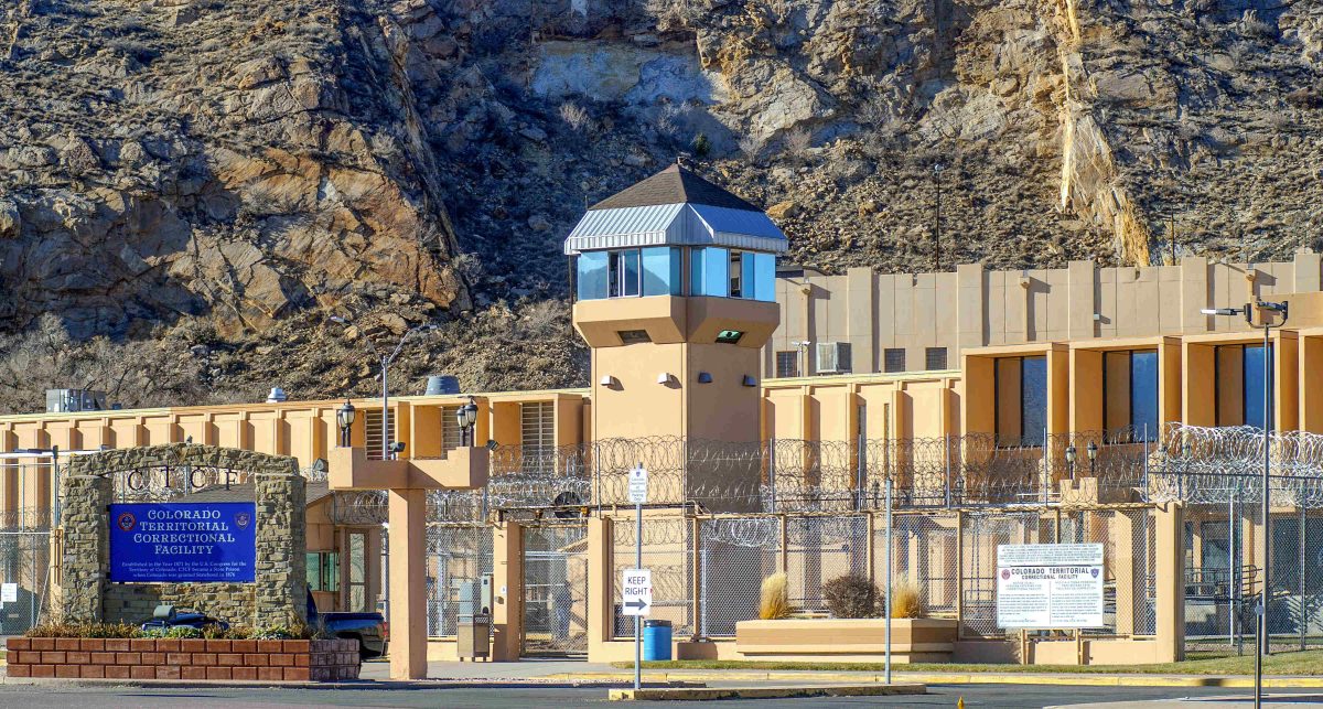 Exterior view of a Colorado correctional facility with a watchtower, barbed wire fence, and signage. A rocky hillside looms in the background.