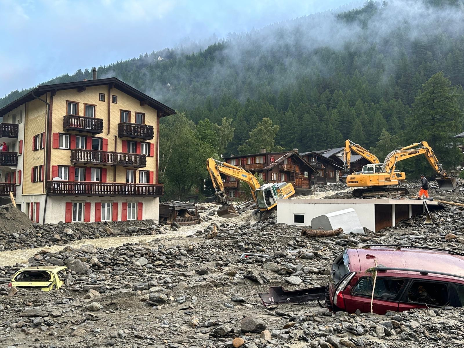 Scene de destruction après une inondation avec des maisons endommagées et des pelleteuses travaillant dans la boue devant des montagnes.