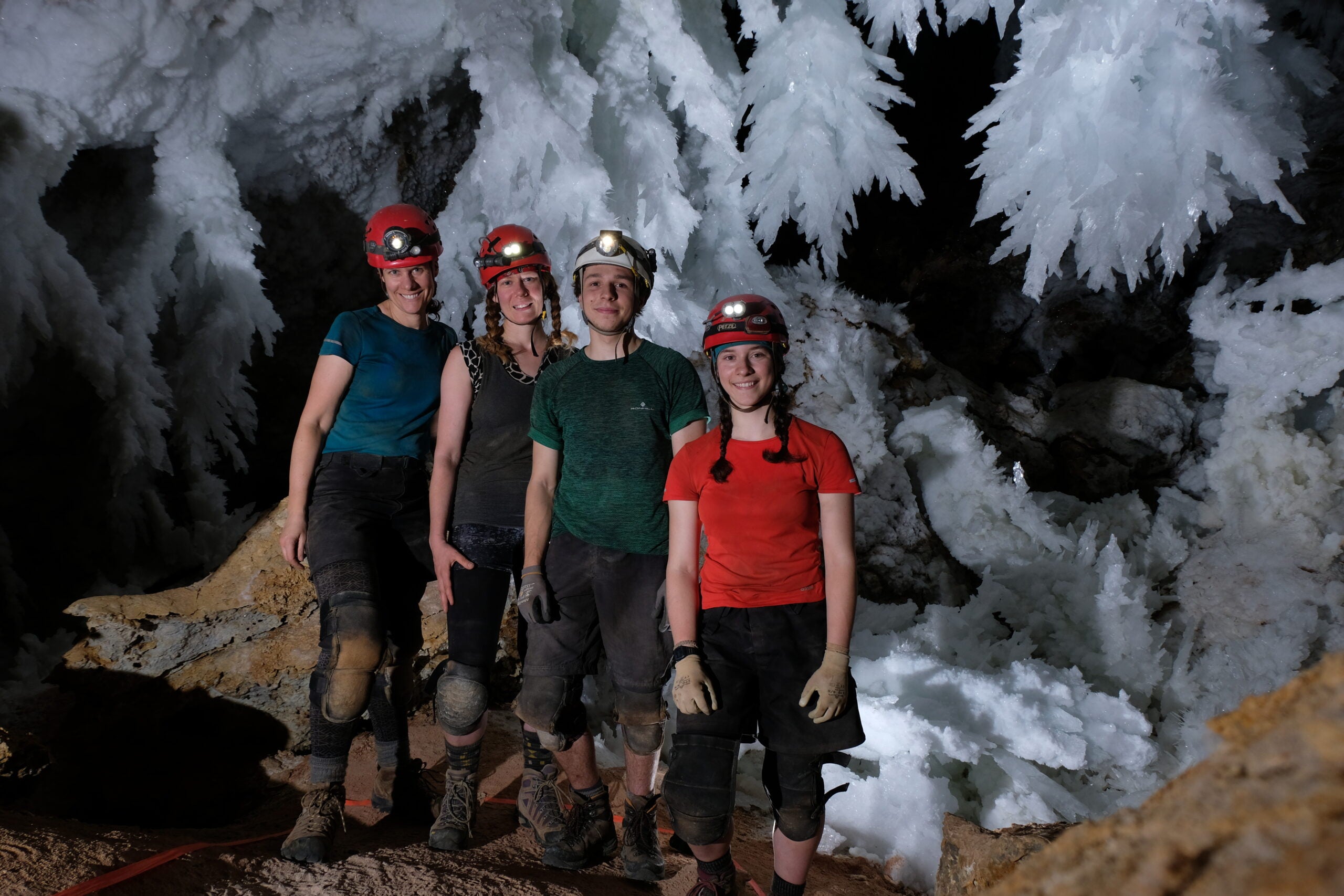 L to R, Hazel Barton, Johanna Kovarik, post-doctoral fellow George Breley, and graduate student Katey Bender in the Chandelier Ballroom of Lechuguilla Cave, New Mexico. Four people wearing helmets with a headlamp stand in front of a huge formation of white crystals. 