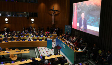 Xi Jinping is visible on a large screen; in the room, dozens of people are seated