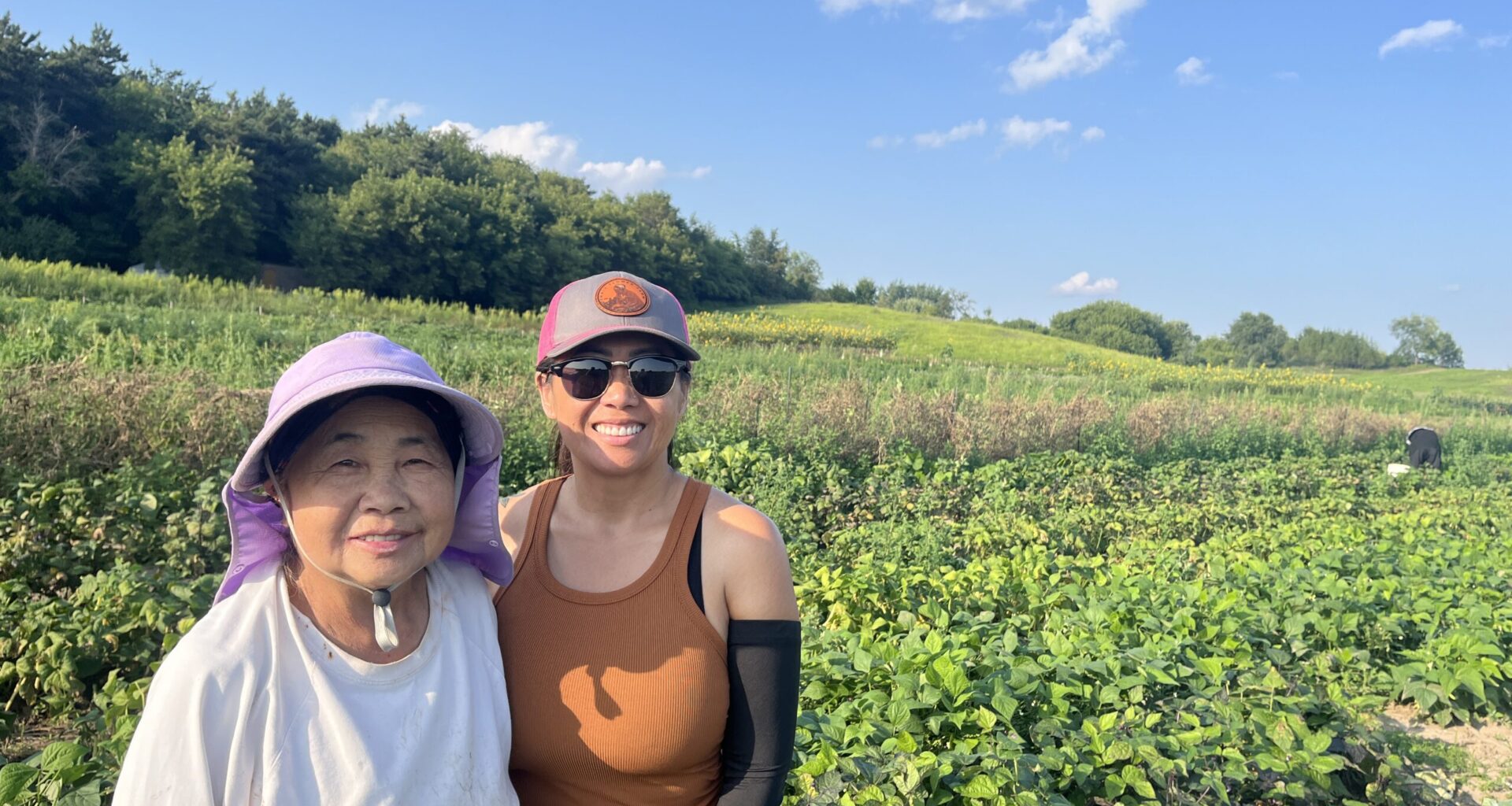 Two women stand in a green field on a sunny day, both smiling at the camera. One wears a sunhat and light shirt; the other wears sunglasses, a cap, and an orange tank top.