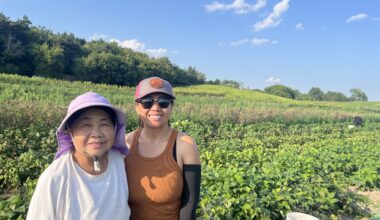 Two women stand in a green field on a sunny day, both smiling at the camera. One wears a sunhat and light shirt; the other wears sunglasses, a cap, and an orange tank top.