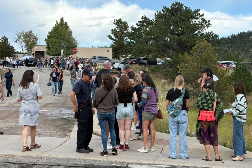 A long line of people stands and waits outside near trees and parked cars on a cloudy day. Some people talk to uniformed officers, while others look ahead or use their phones.