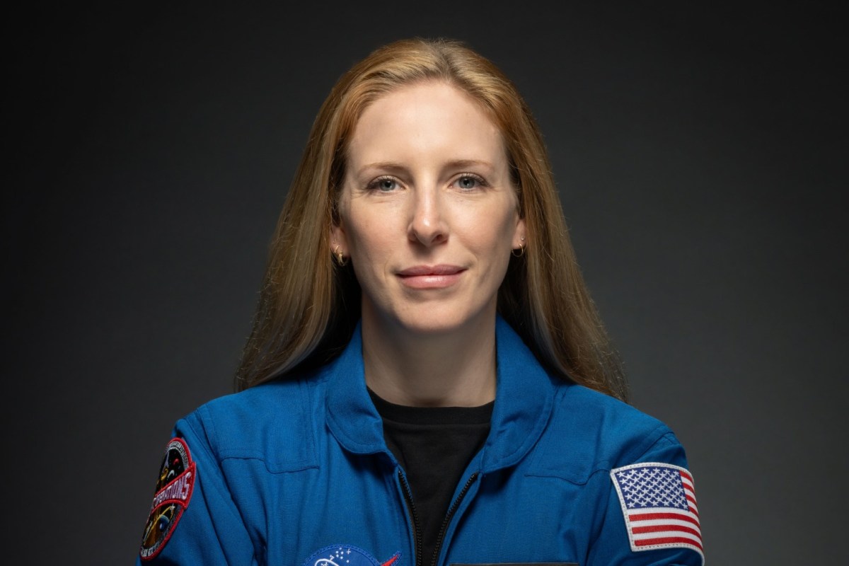 A woman in a blue NASA flight suit with mission patches and an American flag patch poses against a dark background.