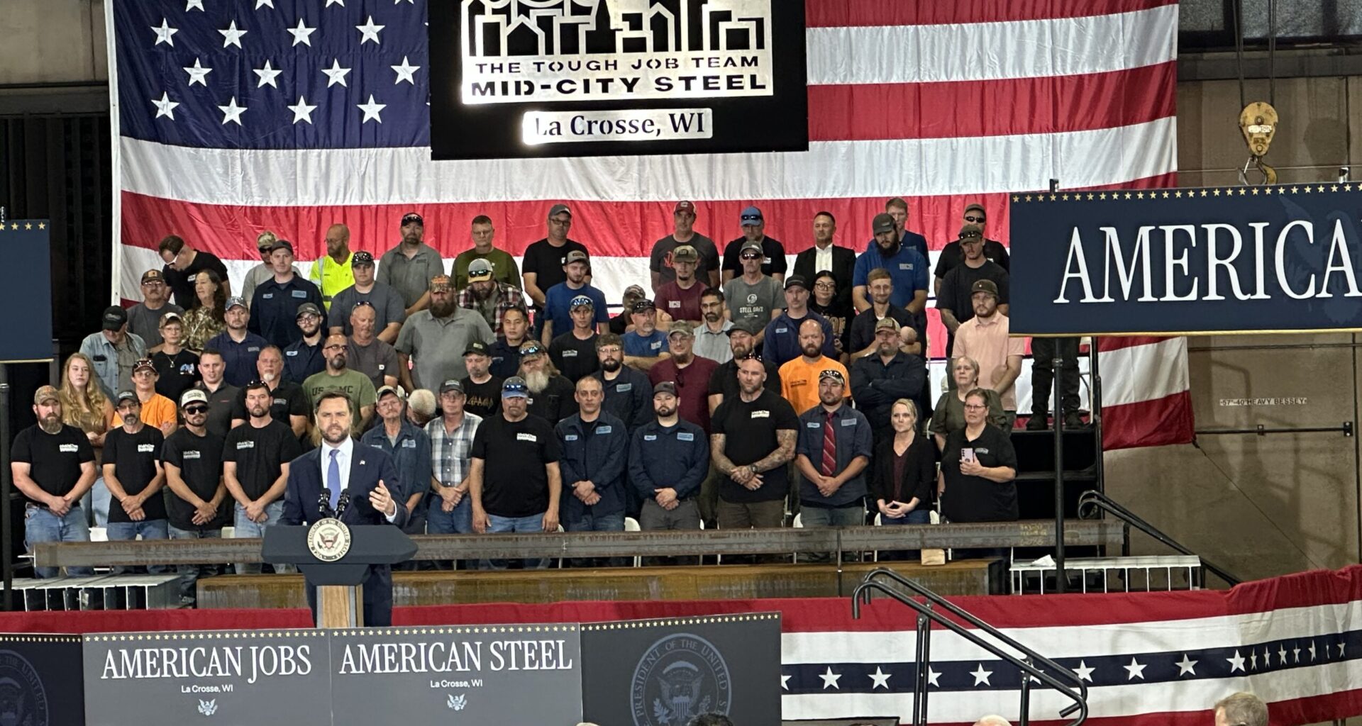 A man speaks at a podium labeled “American Jobs” in front of a large U.S. flag, with a group of people standing behind him and an audience seated in front.