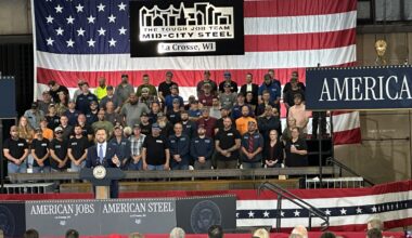 A man speaks at a podium labeled “American Jobs” in front of a large U.S. flag, with a group of people standing behind him and an audience seated in front.
