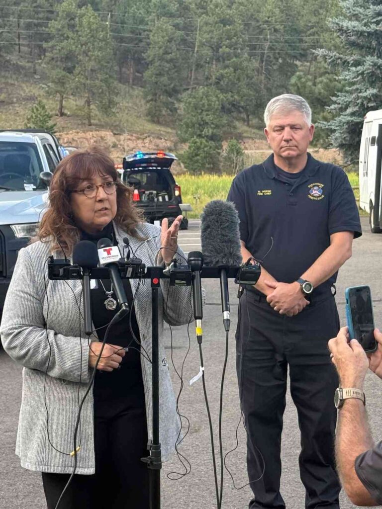 A man and a woman stand in front of a podium with mics in a parking lot with trees in the background