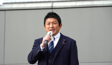 Sohei Kamiya wearing a dark blue suit and tie talking into a microphone.