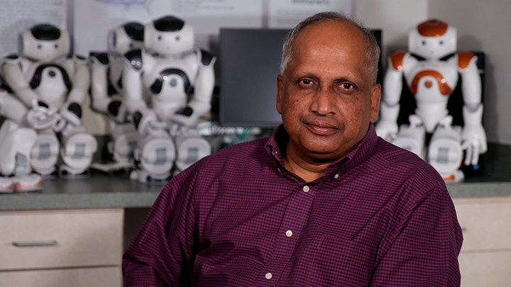 Sam Thangiah sitting in the Robotics Lab at SRU in front of humanoid robots.