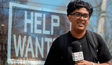 A teen boy in a black shirt with glasses holds a CBC Kids News microphone. Behind him is a help wanted poster.