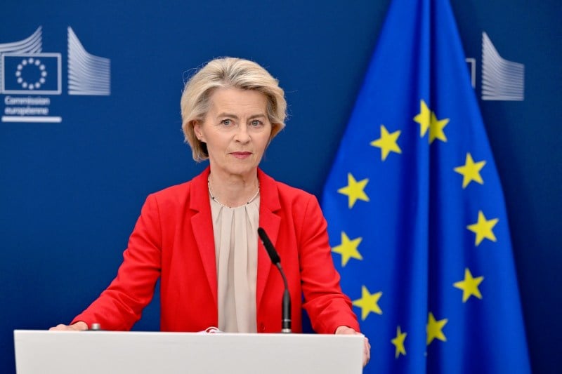 Ursula von der Leyen wears a beige blouse and red blazer while standing behind a podium. Behind her is the European Union flag.