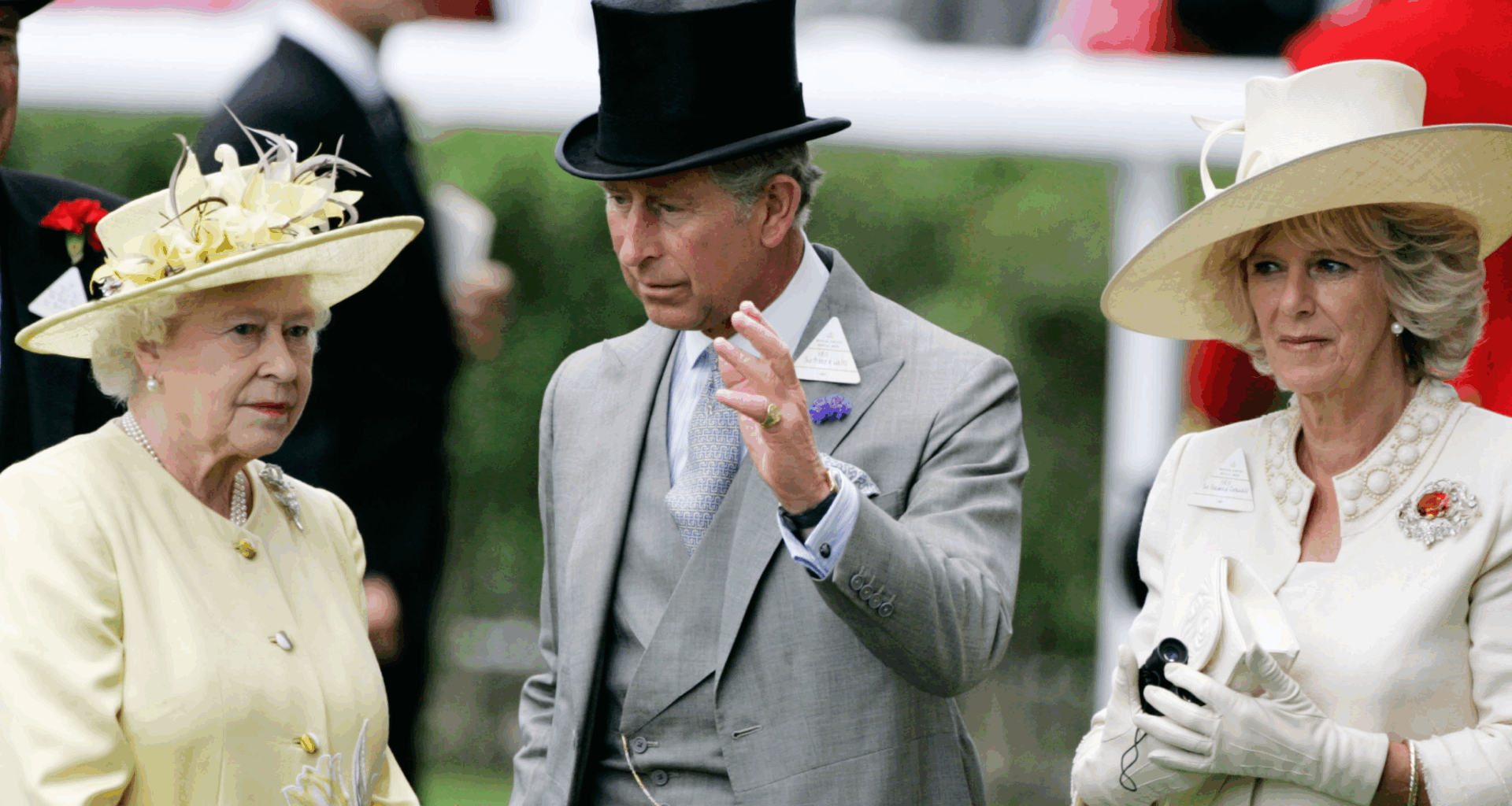 Queen Elizabeth II wears a pale yellow outfit while her son King Charles waves in front of her face, and Queen Camilla stands to the side ignored wearing white