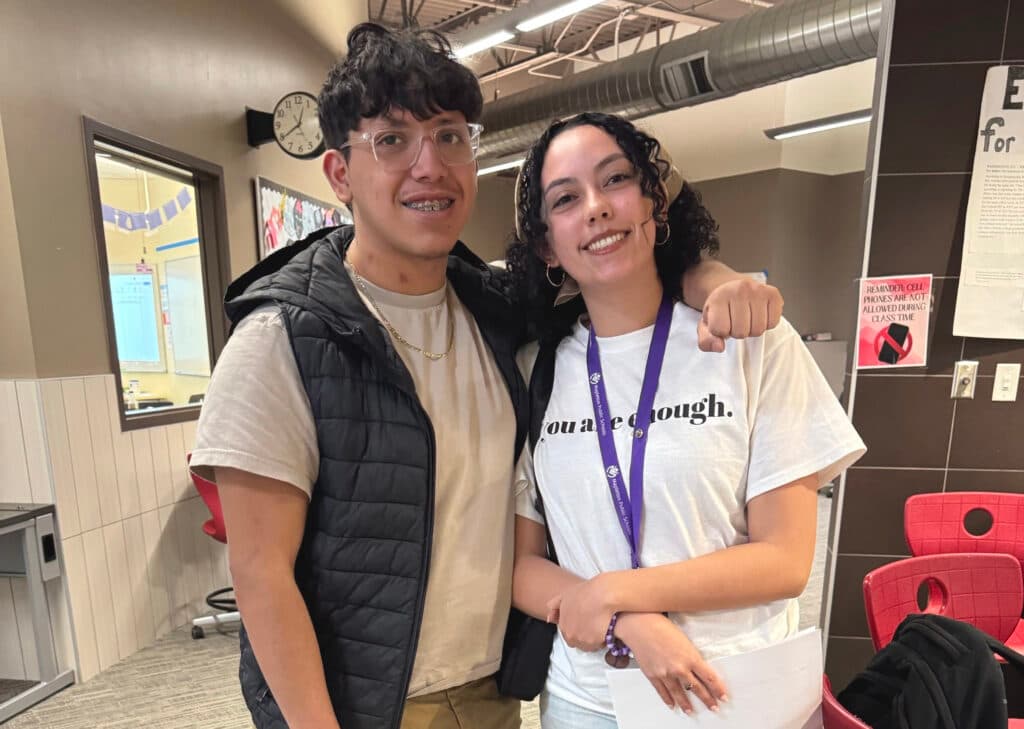 A young boy in clear-framed glasses and braces puts his arm around a woman to his left. She wears a lanyard and a T-shirt that says "You are enough." Both are looking at the camera and smiling.