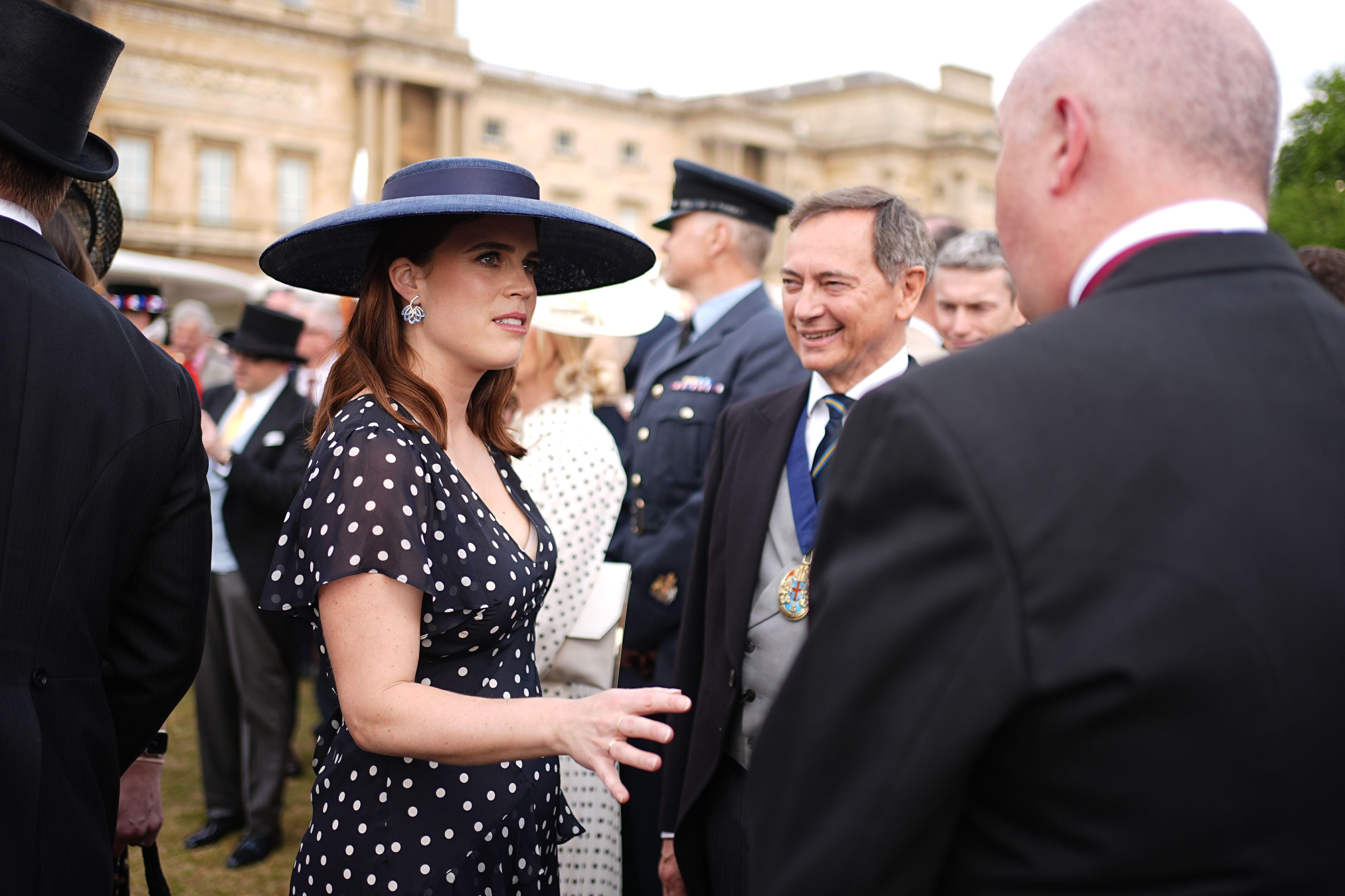 Princess Eugenie wearing a blue polka dot dress and hat talking to men at a garden party