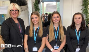 Three young girls standing next to their female college tutor. The tutor has short blonde hair and is wearing glasses with thick black frames. The two girls to her left have long blonde hair. The girl on the end of the line has long dark hair. All of them are wearing black and have lanyards around their necks.