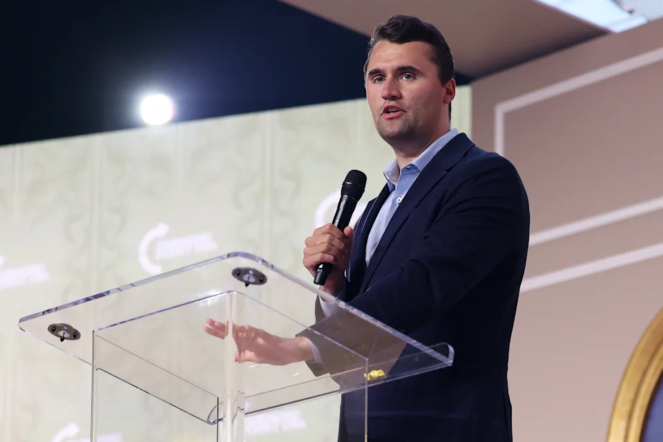Charlie Kirk in a suit speaking at a podium with a microphone, in a formal event setting