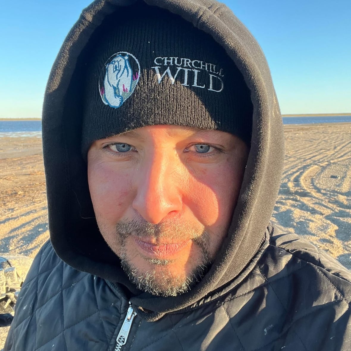 A Cree man with blue eyes, and goatee, wearing a hat and hood stares intently into the camera.