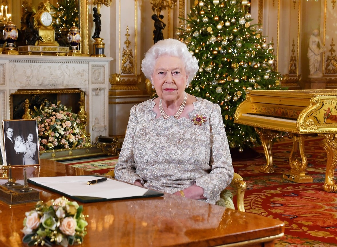A person sits behind a desk in front of a Christmas tree and fireplace.