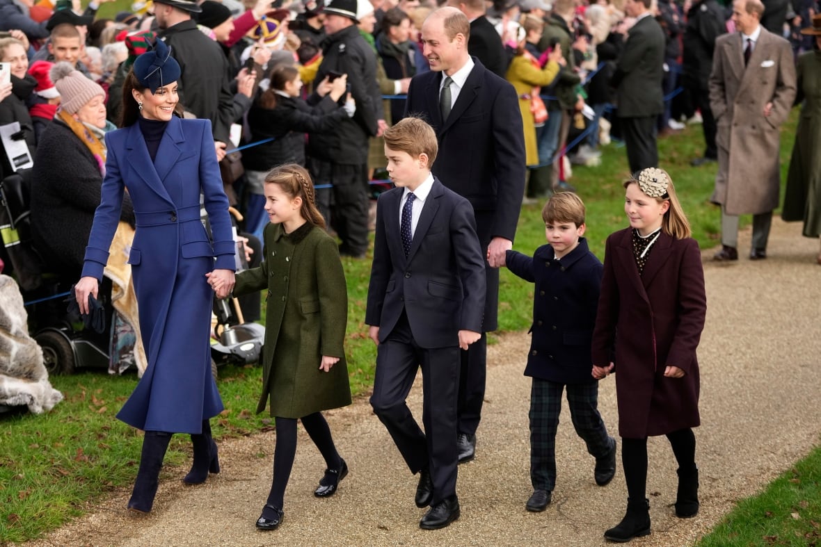 Two adults and four children walk along a gravel walk as a crowd watches behind a rope.