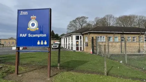 PA Media A blue RAF Scampton sign outside the former air base. Some of the brick buildings can be seen behind the fence. There is grass and a white car parked in the distance.