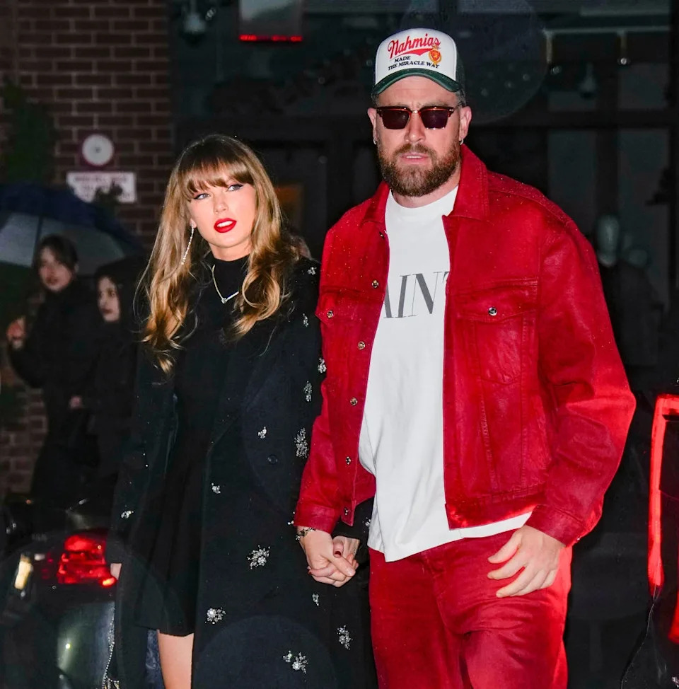 Two people walk hand-in-hand; one in a black outfit with a coat, the other in a red jacket, shirt, pants, and a baseball cap