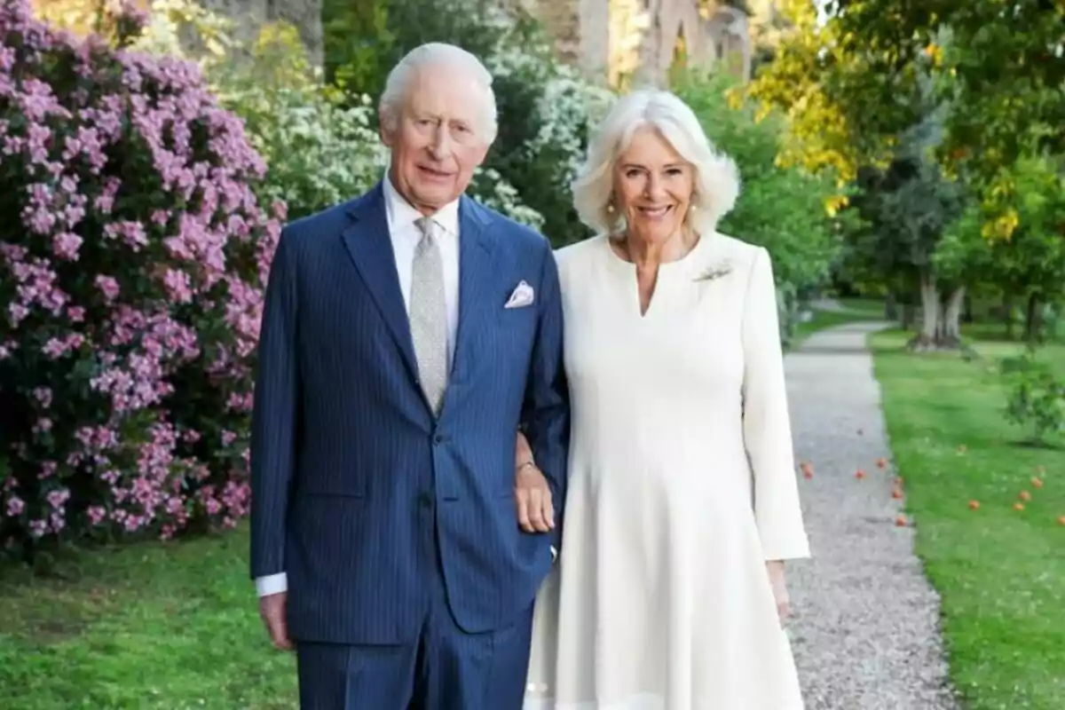 An elderly couple smiles as they walk through a garden with flowers and trees around them.