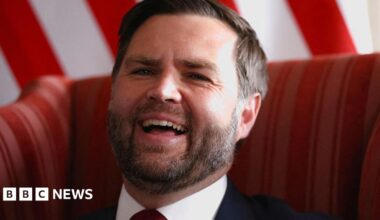 Dolly Mavies in a dark red jacket standing in a field holding a guitar