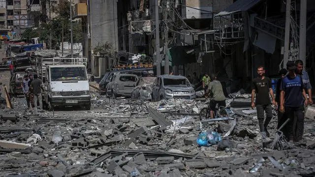Una calle en el vencindario de Rimal y la Ciudad de Gaza está cubierta de escombros. Se ven muchos autos destruidos al lado de la calle, con gente congregándose en pequeños grupos  
