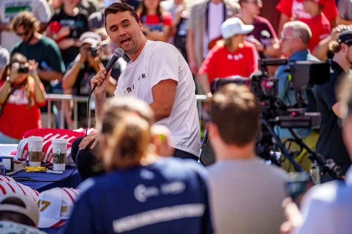 Charlie Kirk speaking into a microphone at an outdoor event, with spectators and camera operators in the background