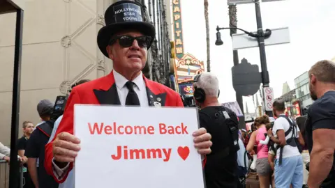 Reuters A man in a red suit and black top hat holding a sign saying "Welcome back Jimmy"
