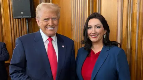 NI Executive/PA Media Donald Trump meets Deputy First Minister Emma Little-Pengelly. She is wearing a red dress with a blue blazer. He is wearing a navy suit with a red tie. The pair are both smiling while standing in a wooden panelled room.